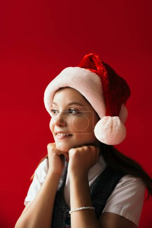 lächelndes und verträumtes Mädchen mit Weihnachtsmütze, das die Hände am Kinn hält und isoliert auf rotem Grund wegschaut
