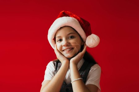 smiling girl in santa hat and beaded bracelet holding hands near face isolated on red