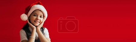 fröhliches Mädchen mit Weihnachtsmannmütze, das in die Kamera lächelt, während es die Hände in der Nähe des Gesichts hält, isoliert auf rotem Banner