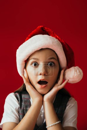 portrait of amazed girl in santa hat looking at camera and holding hands near face isolated on red