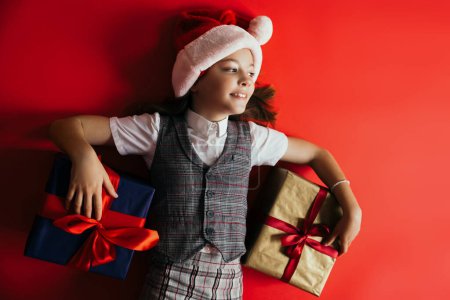 top view of joyful girl in santa hat and checkered vest lying near gifts on red background