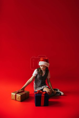 full length of preteen girl in santa hat and gumshoes sitting near gift boxes on red background with copy space