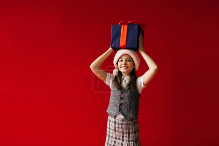 fröhliches Mädchen in karierten Kleidern und Weihnachtsmütze mit Weihnachtsgeschenken über dem Kopf isoliert auf rot