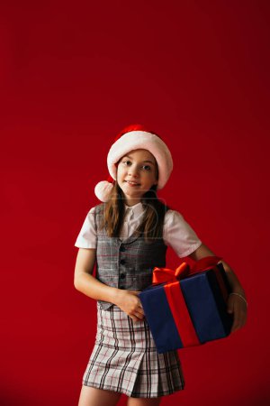 smiling child in santa hat and plaid skirt standing with Christmas present isolated on red