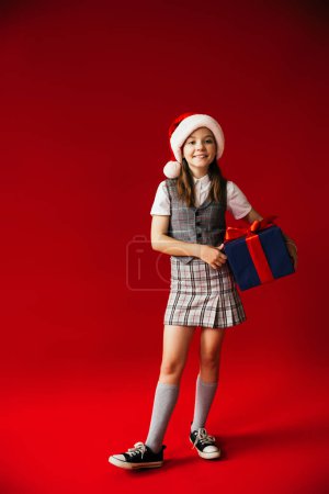 volle Länge des Mädchens in Gummischuhen und kariertem Rock trägt Weihnachtsmann Hut und hält blaue Geschenkbox auf rotem Hintergrund