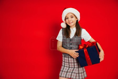 preteen girl in stylish checkered clothes and santa hat holding Christmas present isolated on red