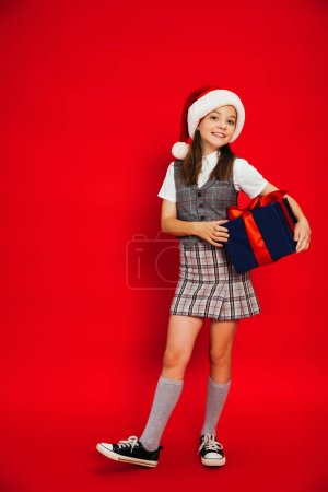volle Länge des fröhlichen Kindes in kariertem Rock und Weihnachtsmütze mit blauem Geschenkkarton auf rotem Hintergrund
