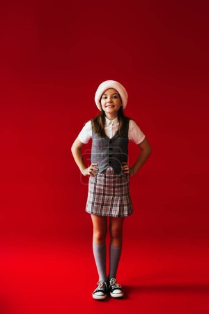 full length of girl in checkered skirt and santa hat posing with hands on hips on red background