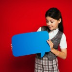 Smiling preteen girl in plaid skirt looking at blue speech bubble isolated on red