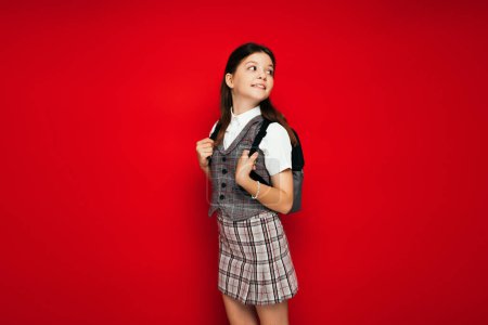 smiling girl in plaid clothes standing with backpack and looking back isolated on red, banner 