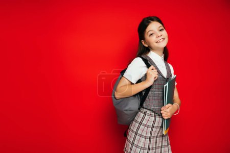 stylisches Schulmädchen mit Schulheften und Rucksack, das isoliert auf rotem Banner in die Kamera lächelt 