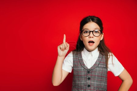 Photo for Astonished girl in eyeglasses showing idea gesture and looking at camera isolated on red - Royalty Free Image