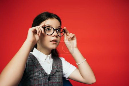 girl in plaid vest looking away and adjusting eyeglasses isolated on red