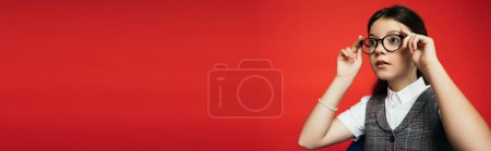 brunette girl adjusting stylish eyeglasses and looking away isolated on red, banner