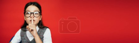 Téléchargez les photos : Brunette preteen fille dans des lunettes montrant le silence geste isolé sur rouge, bannière - en image libre de droit