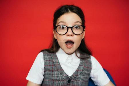 astonished girl in eyeglasses and plaid vest looking at camera isolated on red