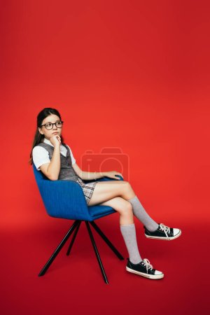 volle Länge des nachdenklichen Mädchens in Gummischuhen und Brille im Sessel sitzend und auf rotem Hintergrund wegschauend