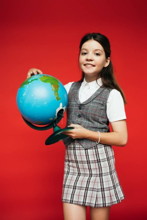 smiling girl in plaid vest and skirt holding globe isolated on red, banner 