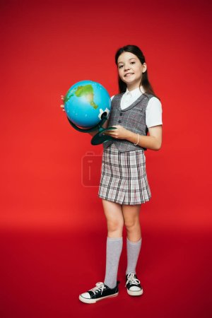 volle Länge des glücklichen Mädchens in kariertem Rock und Gummischuhen posiert mit Globus auf rotem Hintergrund