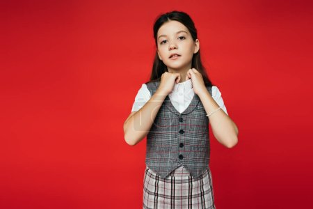 preteen girl in checkered vest and skirt adjusting collar of white blouse isolated on red, banner 