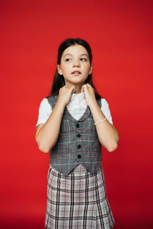 preteen schoolgirl in checkered skirt and vest adjusting collar of blouse and looking away isolated on red, banner 
