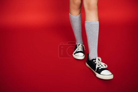 Téléchargez les photos : Vue partielle de l'enfant en chaussettes de genou grises portant des gommes noires et blanches sur fond rouge - en image libre de droit