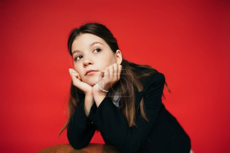 portrait of thoughtful brunette girl in black blazer holding hands near face and looking away isolated on red