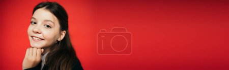Foto de Retrato de chica morena sonriente con la mano cerca de la barbilla mirando a la cámara aislada en rojo, pancarta - Imagen libre de derechos