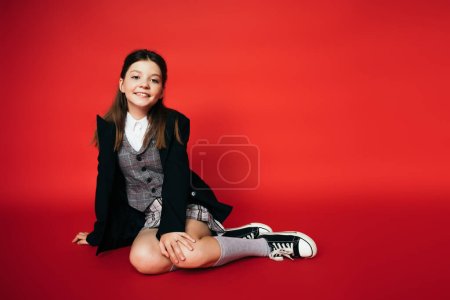 full length of pleased girl in black blazer and gumshoes sitting on red background