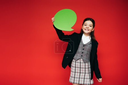 happy girl in black jacket and plaid skirt holding green speech bubble isolated on red