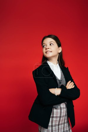 happy girl in checkered skirt and black blazer standing with crossed arms and looking back isolated on red