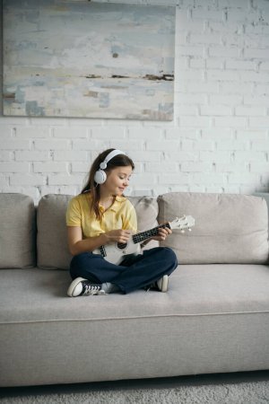 Positives Kind mit Kopfhörern, das mit überkreuzten Beinen auf einer modernen Couch sitzt und Ukulele spielt
