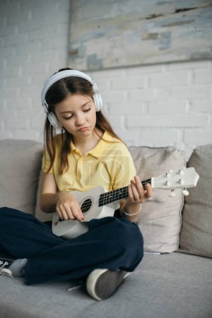 unzufriedenes Mädchen mit Kopfhörern, das kleine hawaiianische Gitarre spielt, während es mit überkreuzten Beinen auf der Couch sitzt