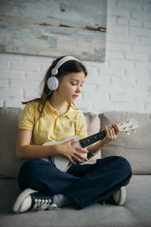 preteen girl in wired headphones playing ukulele while sitting with crossed legs on couch 
