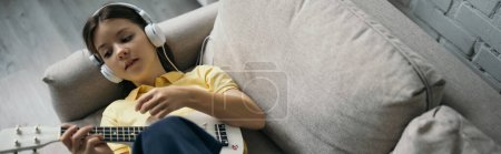 top view of preteen girl in wired headphones lying on couch and playing ukulele, banner