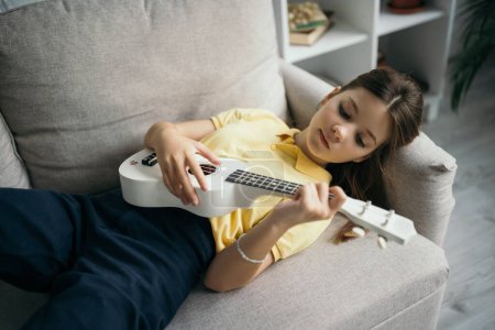 Téléchargez les photos : Vue grand angle de preteen fille couché sur canapé à la maison et jouer petite guitare hawaïenne - en image libre de droit