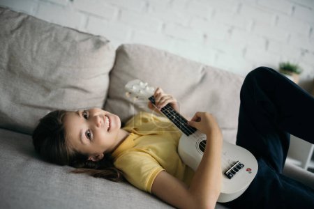Photo for Cheerful girl looking at camera while playing small hawaiian guitar on sofa at home - Royalty Free Image