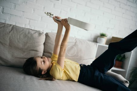 niña preadolescente acostada en el sofá en la sala de estar y la celebración de ukelele en manos levantadas