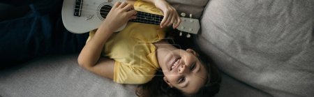 Draufsicht des fröhlichen Mädchens, das auf der Couch liegt und kleine hawaiianische Gitarre spielt, während es in die Kamera schaut, Banner