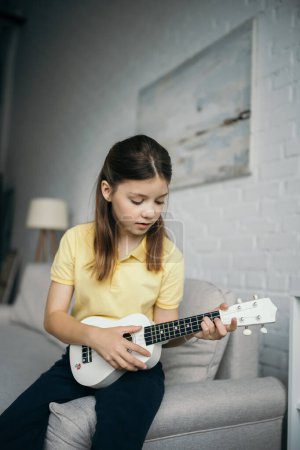preteen girl sitting on couch in blurred living room and playing ukulele