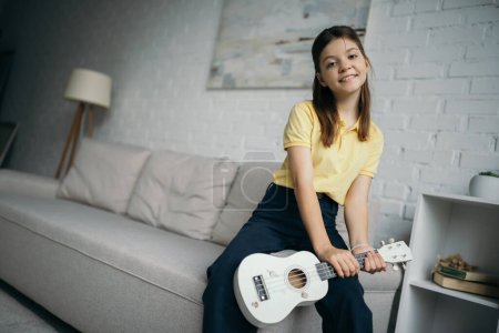 happy preteen girl holding ukulele and looking at camera near blurred couch at home