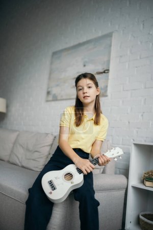 chica preadolescente con pequeña guitarra hawaiana mirando a la cámara cerca del sofá en la sala de estar