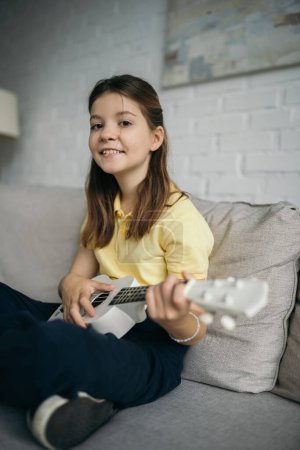 cheerful preteen girl smiling at camera while playing ukulele on sofa in living room