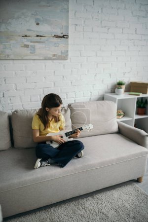 Téléchargez les photos : Vue grand angle de la fille assise sur le canapé moderne avec les jambes croisées et jouer ukulele - en image libre de droit