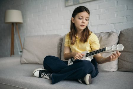 Photo for Brunette girl sitting on couch with crossed legs and tuning small hawaiian guitar - Royalty Free Image