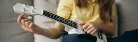 Téléchargez les photos : Vue recadrée de l'enfant flou accordant la petite guitare hawaïenne à la maison, bannière - en image libre de droit