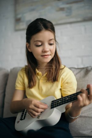 sonriente chica jugando pequeña guitarra hawaiana en casa sobre fondo borroso