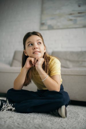 Photo for Smiling and dreamy girl sitting on floor with crossed legs and looking away - Royalty Free Image