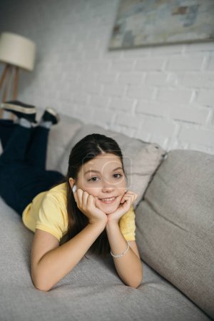 fröhliches Mädchen, das im drahtlosen Kopfhörer Musik hört, während es mit den Händen in der Nähe des Gesichts auf der Couch liegt