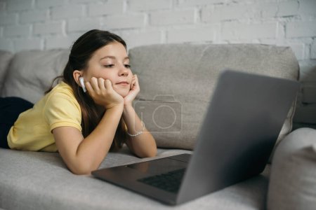 brunette girl in wireless earphone looking at blurred laptop while lying on cozy couch at home